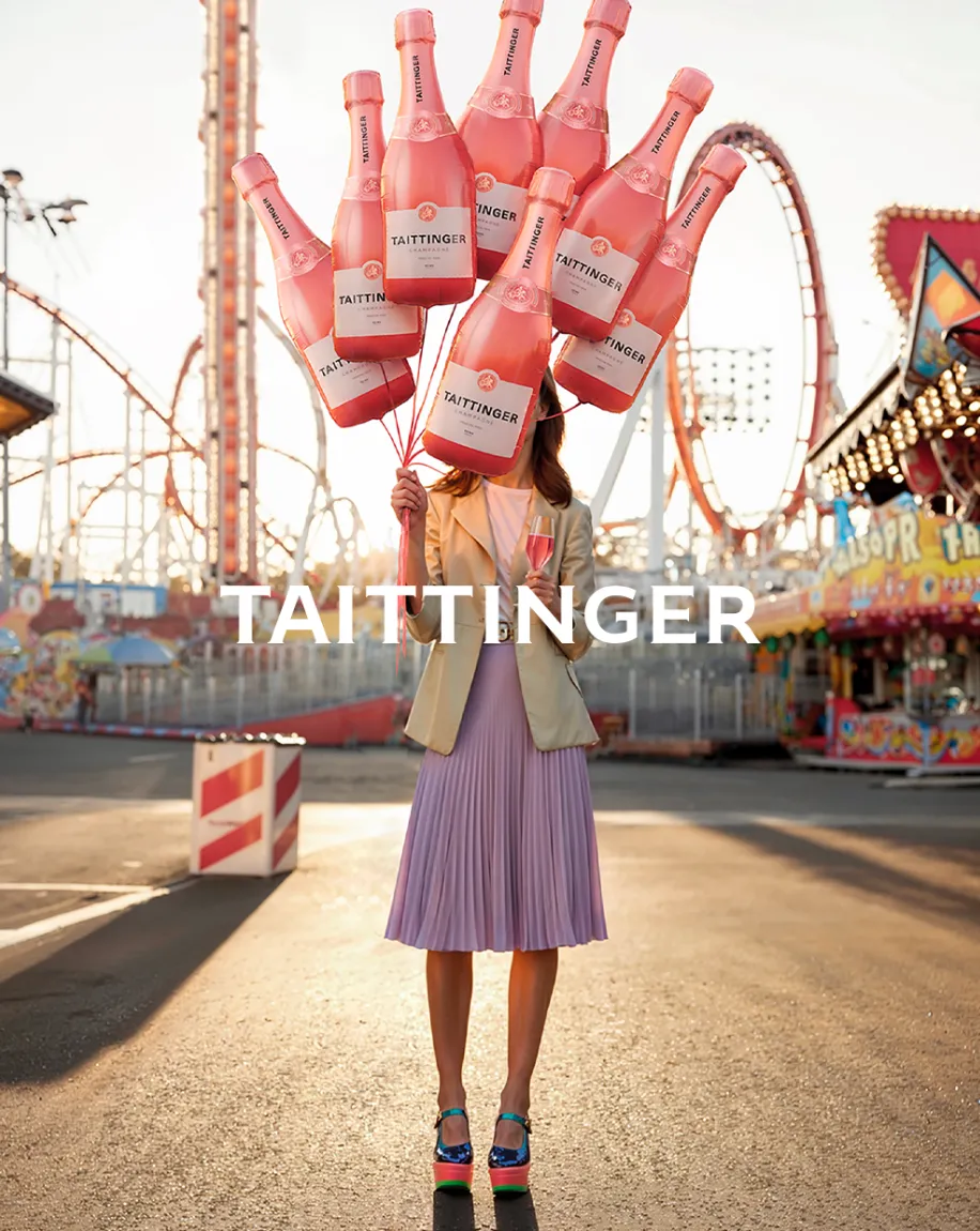 Woman at a fairground holding a bouquet of giant pink Taittinger champagne bottle balloons against roller coasters — cgi studio Thinkdiff, photography & art direction by Floow, produced by Thinkdiff AI Production House