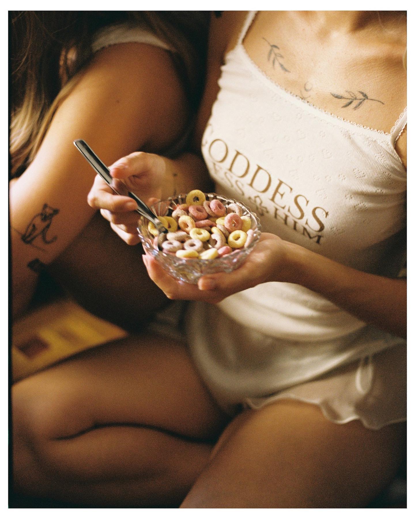 Woman in a cream slip dress eating from a bowl, seated cross-legged with a visible arm tattoo, warm film-toned lighting — ai advertising agency Thinkdiff, generative AI artwork & art direction by Noah Pharrell