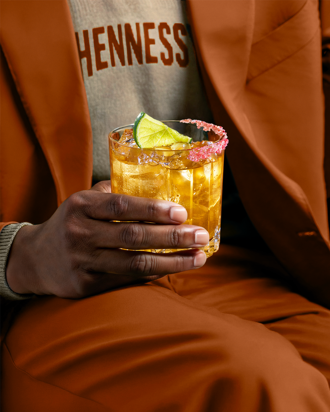 Close-up of a hand holding an orange cocktail with a lime garnish, warm suit fabric in frame — ai video production Thinkdiff, photography & art direction by Thinkdiff AI Production House