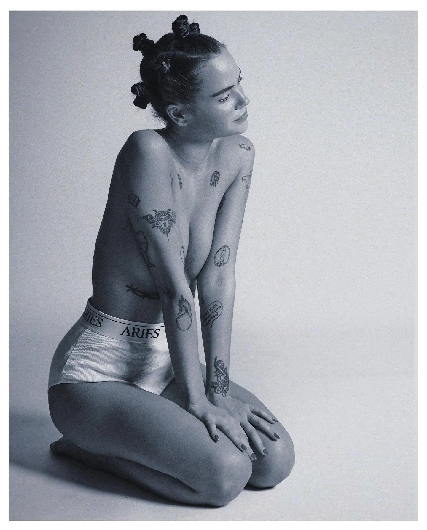 Woman with braided hair and visible tattoos sitting curled on the floor in a minimalist grey studio setting — ai advertising agency Thinkdiff, generative AI artwork & art direction by Noah Pharrell